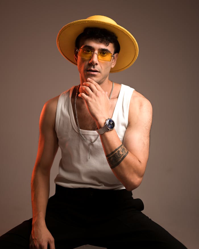 A fashionable man poses in a studio wearing a yellow hat, sunglasses, and modern attire.